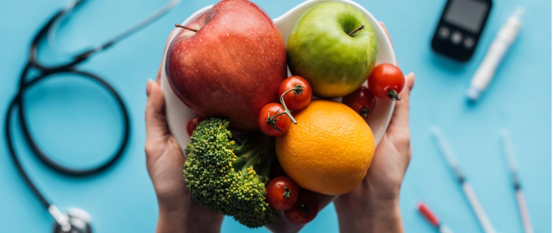 Na zdjęciu w centrum jest widoczna miska trzymana w dłoniach a w niej owoce i warzywa. W tle widać stetoskop, strzykawki i glukometr. 