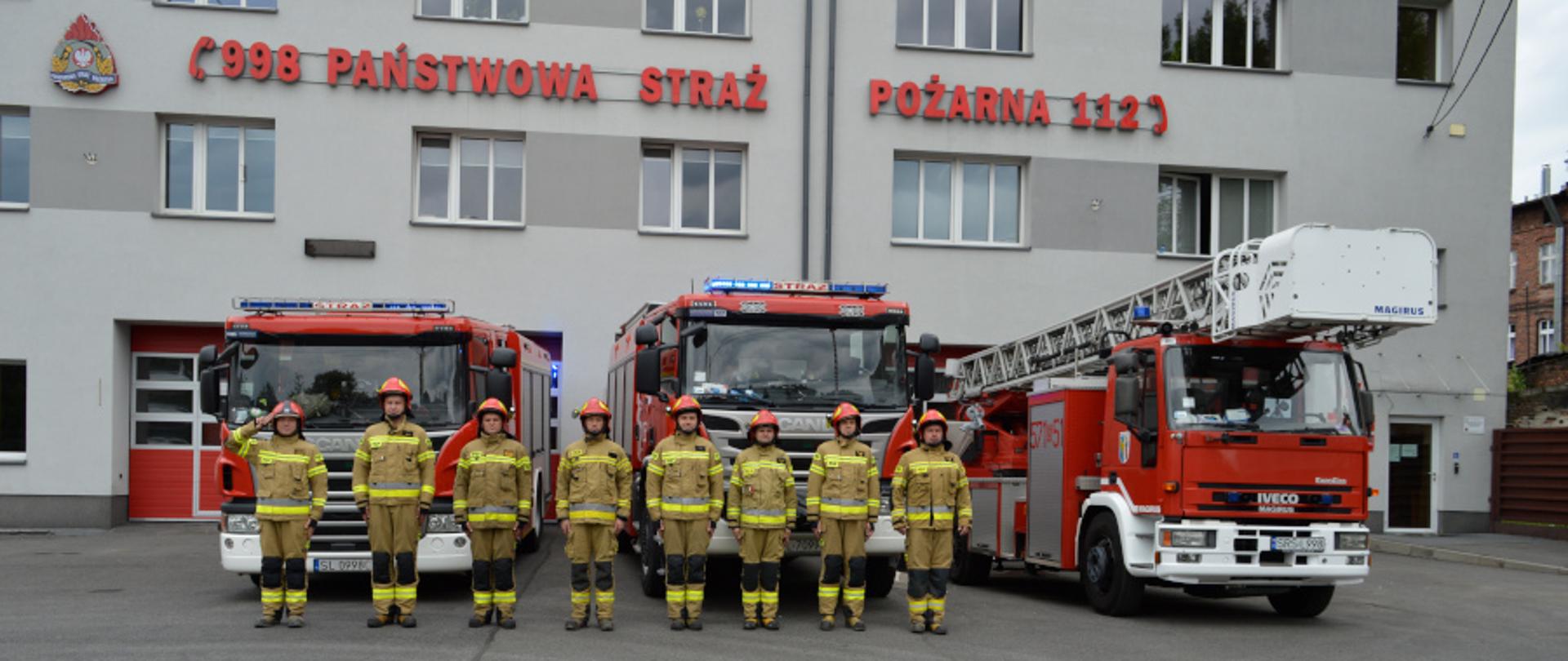 Na tle budynku Komendy Miejskiej Państwowej Straży Pożarnej znajdują się samochody bojowe, a przed nimi strażacy, dowódca pododdziału oddaje honor.