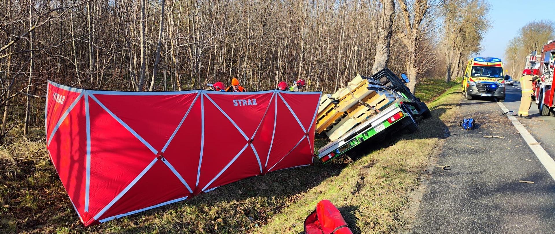 Zdjęcie przedstawia miejsce wypadku: z lewej stoi rozłożony parawan za którym udzielana jest pomoc poszkodowanemu, przy nim samochód, który uderzył w drzewo, a na dalszym planie widać ambulans i samochód gaśniczy.