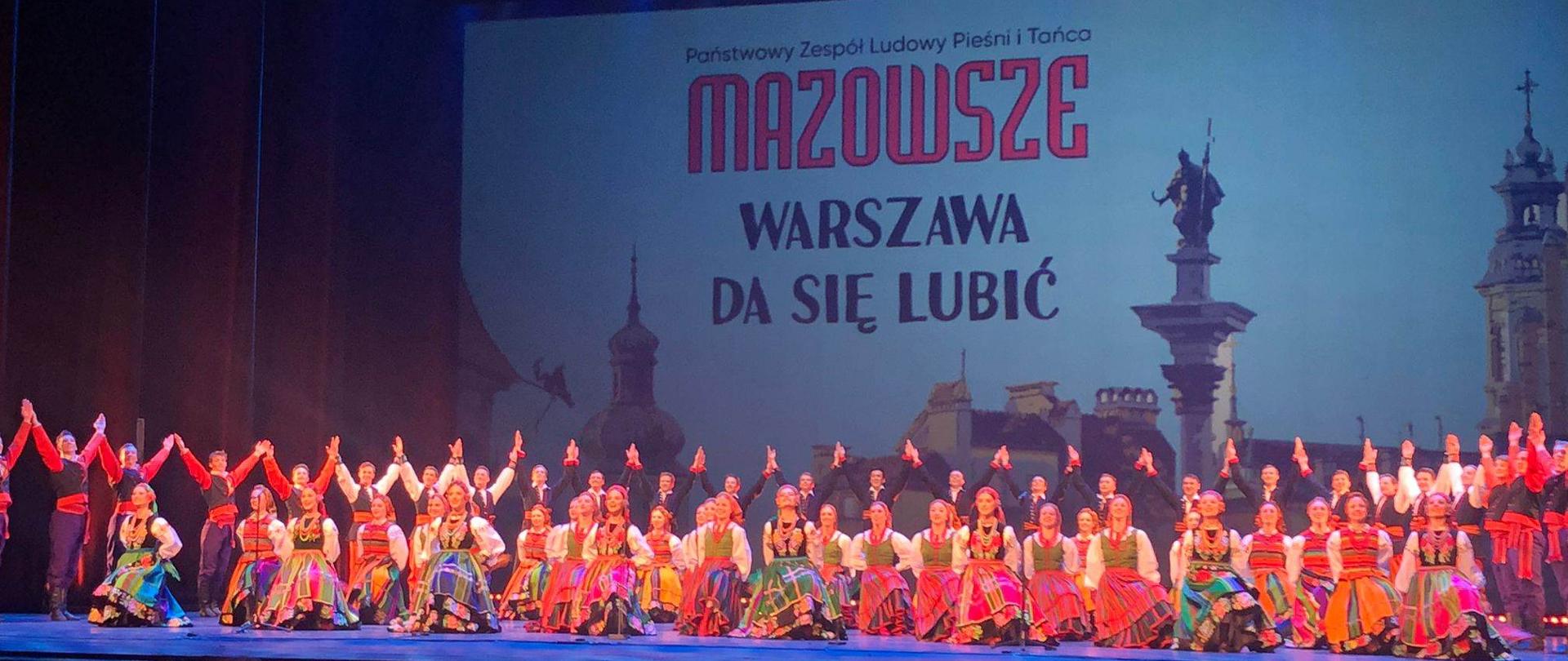 Zdjęcie sceny finałowej występu Państwowego Zespołu Ludowego Pieśni i Tańca Mazowsze w Teatrze Wielkim Operze Narodowej w Warszawie. Tancerki klęczą na jednym kolanie, z tyłu tancerze z podniesionymi rękoma ku górze.