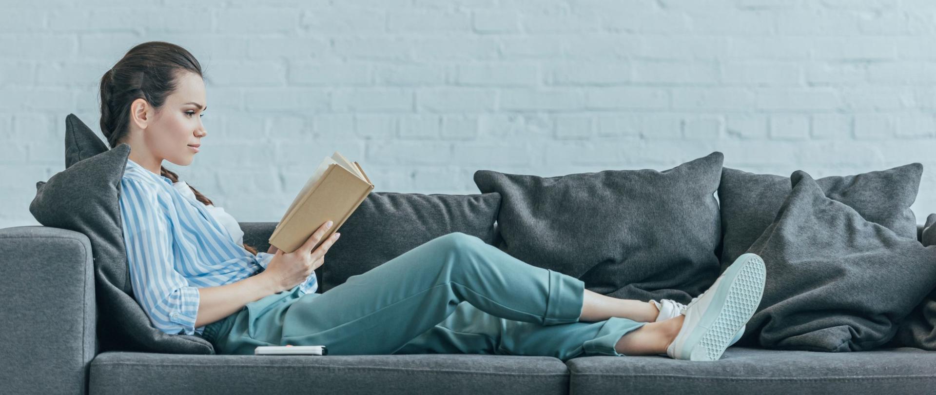 Na zdjęciu jest kobieta leżąca na kanapie, czytająca książkę.