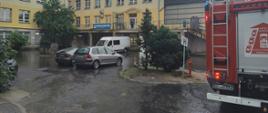 Na zdjęciu widać zaparkowane samochodu przed budynkiem szpitala. Plac jest zalany wodą. Z prawej strony widać samochód strażacki.