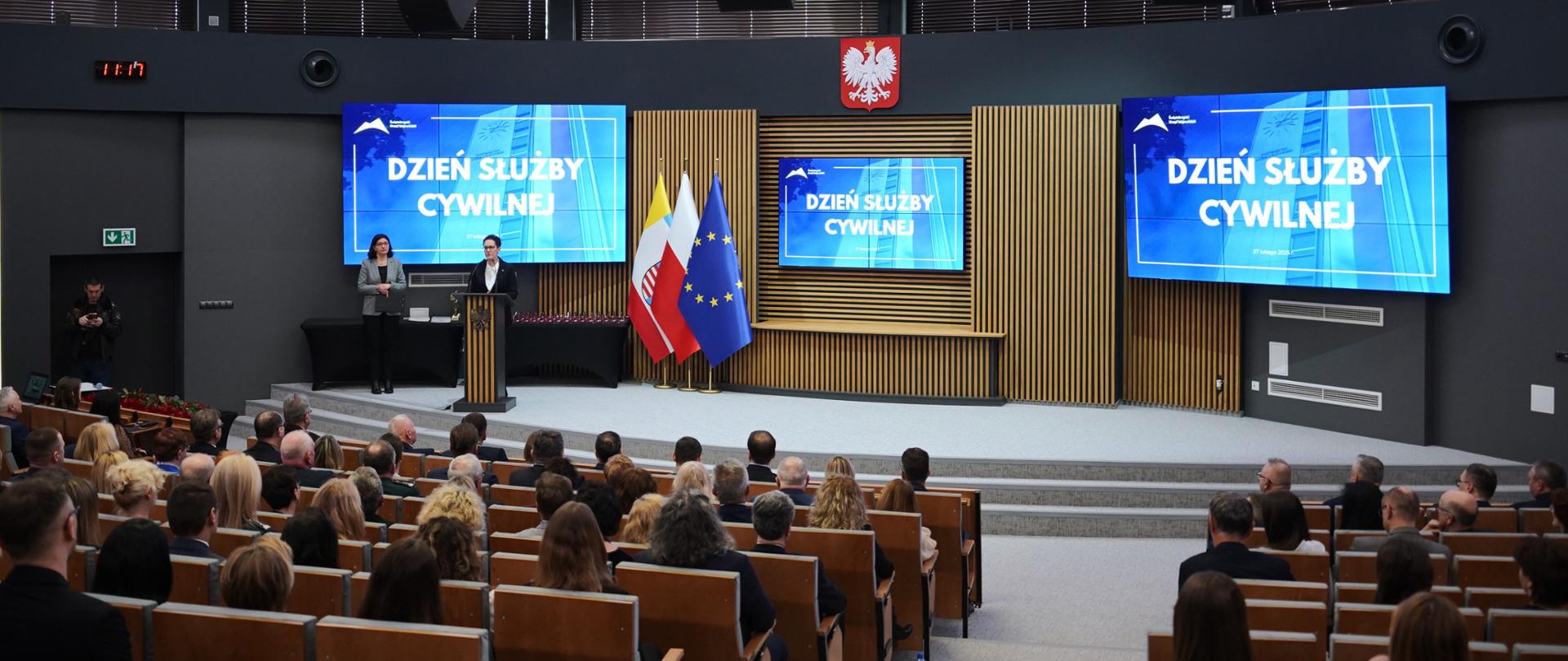 SSC przemawia na podium w sali konferencyjnej ŚUW. W tle ekrany z napisem Dzień Służby Cywilnej. Przed nią siedzą cksc w rzędach.