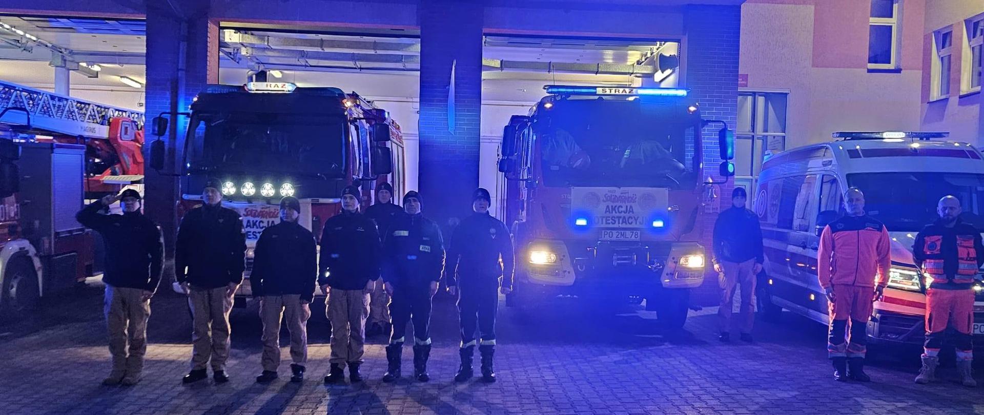 Zdjęcie przedstawia stojących na placu strażaków i ratowników medycznych oddających hołd dla zmarłego ratownika medycznego.
W tle wozy strażackie, ambulans i budynek Komendy.
