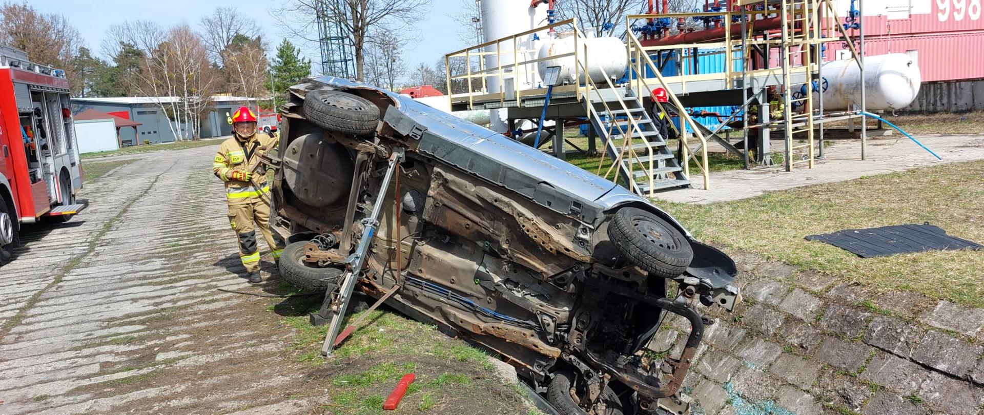 Samochód osobowy wrzucony do rowu na poligonie. Jest ustabilizowany specjalnymi narzędziami. Obok samochodu stoi strażak.