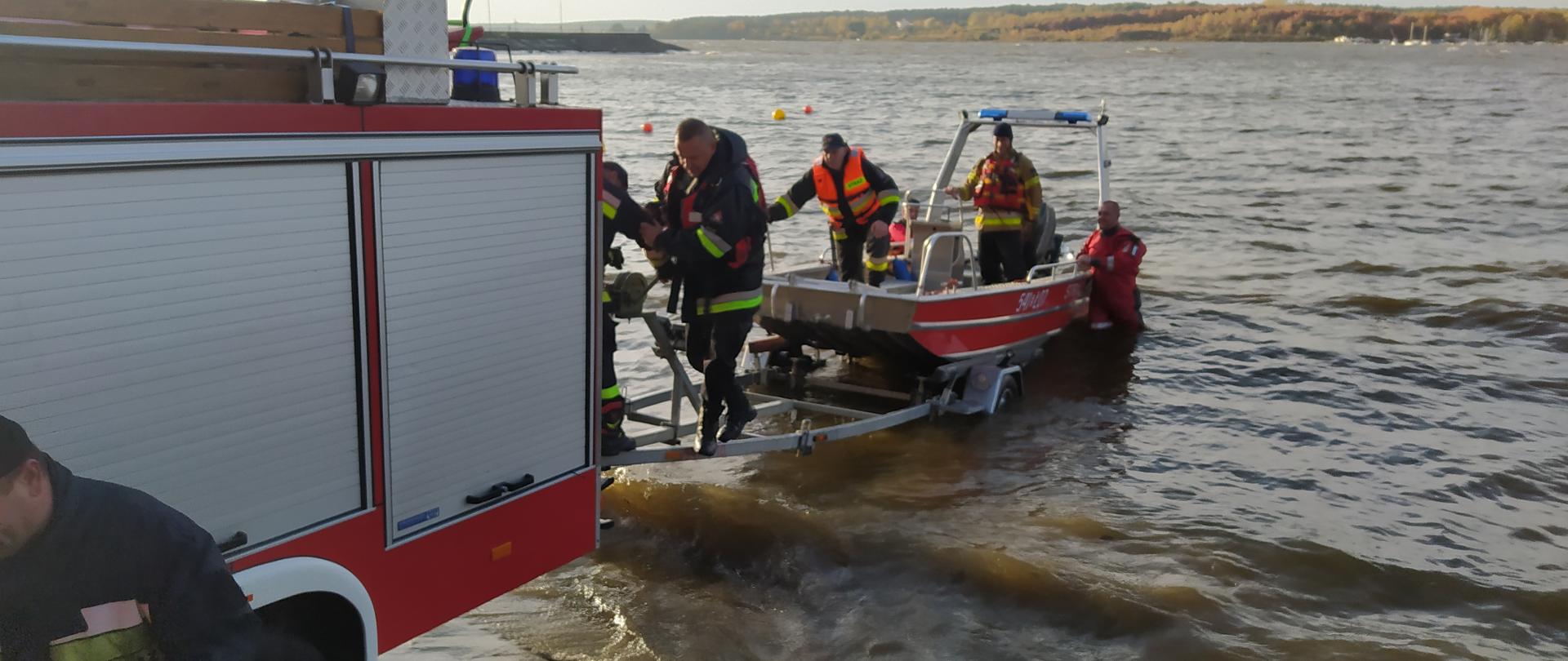 Samochód strażacki stoi nad brzegiem zalewu. Do samochodu przyczepiona jest przyczepka z łódką. Przyczepka jest częściowo w wodzie. Na łódce stoją strażacy. w tle widać dużą taflę wody. 