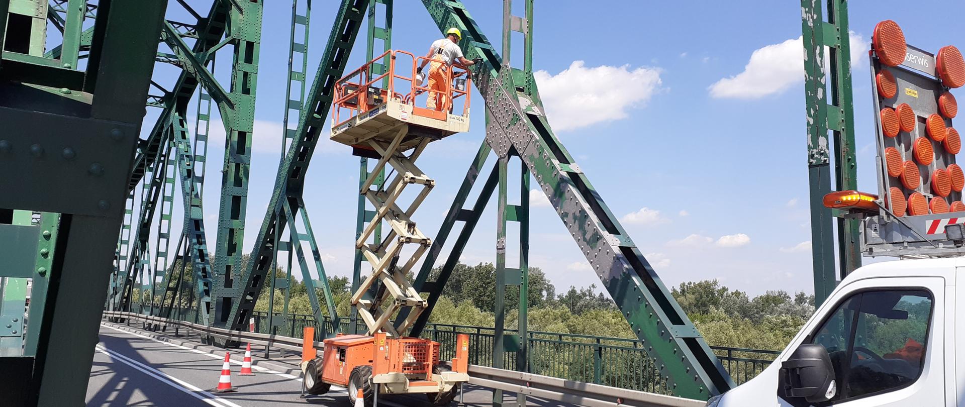 DK73 most w Szczucinie - robotnik stojący na podnośniku maluje zieloną kratownicę mostu. Podnośnik stoi na jezdni. Za nim samochód ze światłami ostrzegawczymi