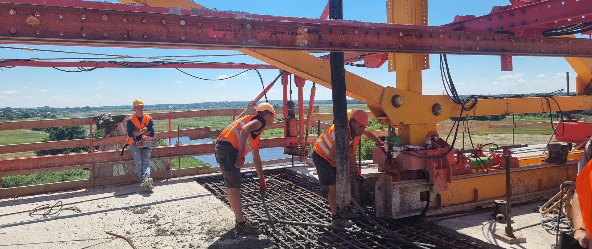 pracownicy budowlani w kaskach i pomarańczowych kamizelkach betonują zazbrojone połączenie mostu nad Narwią. Pracownik dystybuuje beton przy pomocy pompy do betonu, drugi zagęszcza beton zagęszczarką buławową