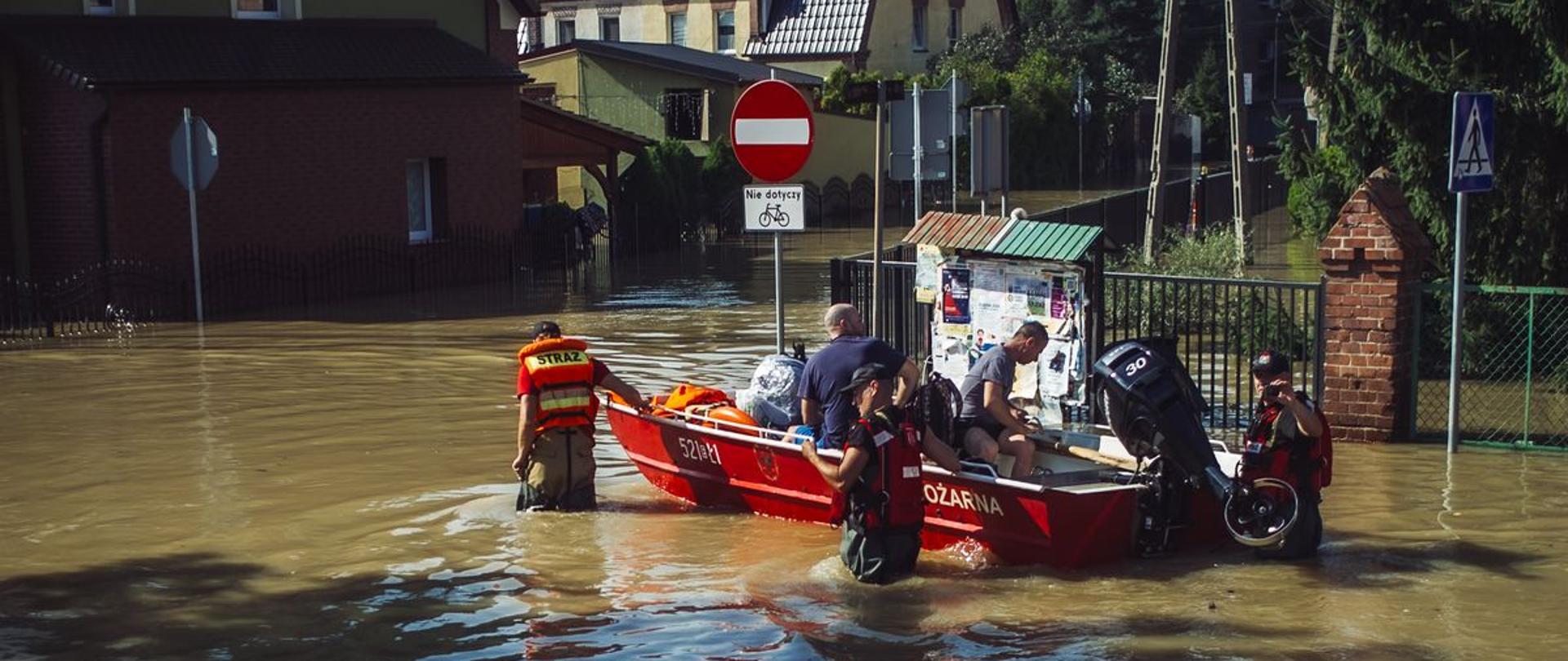 Strażacy Państwowej Straży Pożarnej na łodzi ewakuują kolejne osoby z miejscowości Lewin Brzeski. Strażacy w woderach i kamizelkach asekuracyjnych idą w wodzie przy łodzi.