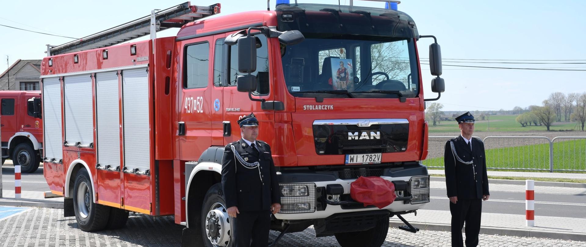 Na zdjęciu znajduje się czerwony samochód ratowniczo-gaśniczy, który został przekazany dla OSP. Obok niego, po obydwu stronach stoją druhowie osp ubrani w ubiór galowy.