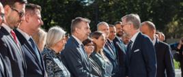 Na dworze w słońcu minister Wieczorek stoi wśród grupy ludzi, podaje rękę stojącemu naprzeciwko mężczyźnie w garniturze, w tle zielone drzewa.