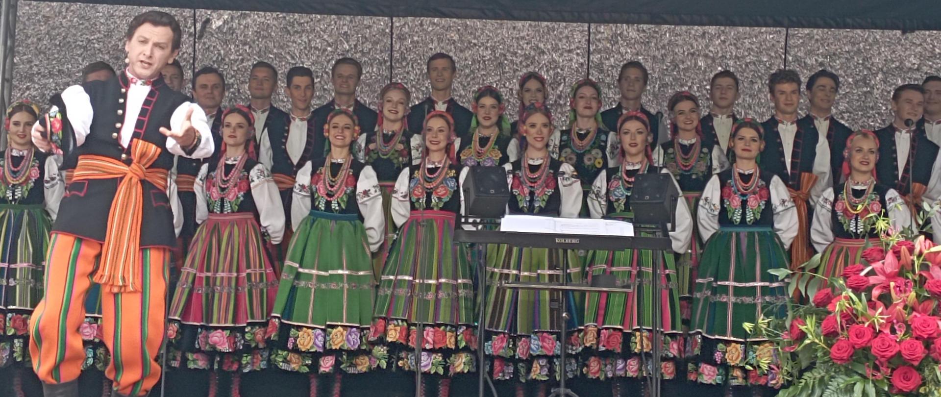 Na scenie stoi grupa mężczyzn i kobiet ubranych w kolorowe stroje ludowe - Państwowy Zespół Ludowy Pieśni i Tańca „Mazowsze”

