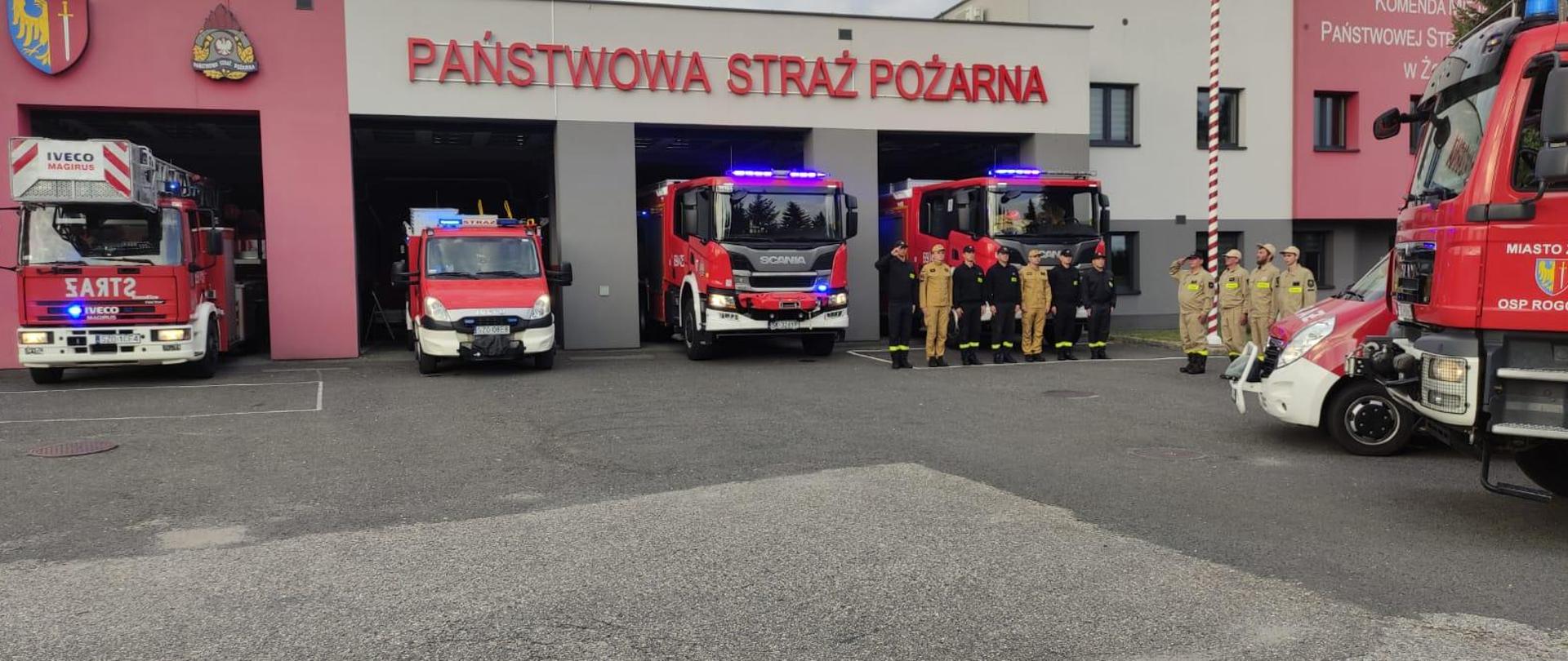 Kilku strażaków stoi ustawionych w szeregu przed samochodami pożarniczymi, dwóch z nich salutuje. Samochody stoją w bramach garażowych, mają włączone światła błyskowe. Nad nimi logo PSP i Miasta Żory oraz napis Państwowa Straż Pożarna.