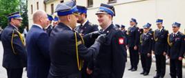 Widok z boku. Zastępca Komendanta Wojewódzkiego PSP w trakcie dekoracji odznaczonych funkcjonariuszy. 
