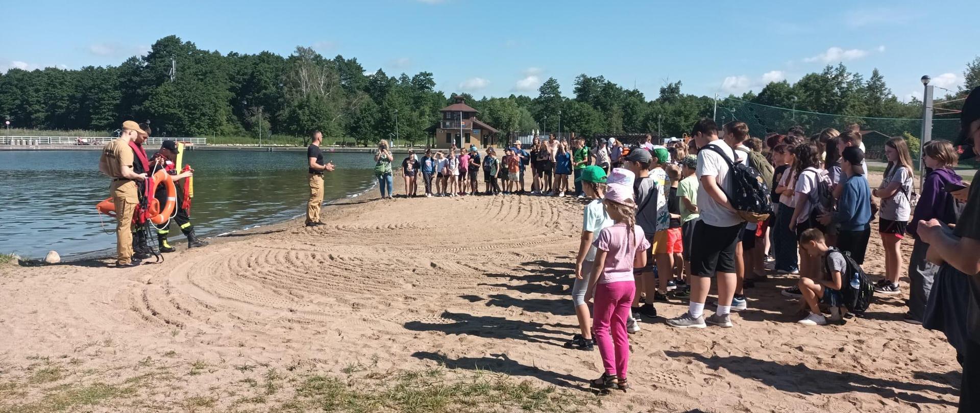 Strażacy stoją przed dziećmi na plaży i omawiają zasady postępowania nad wodą.