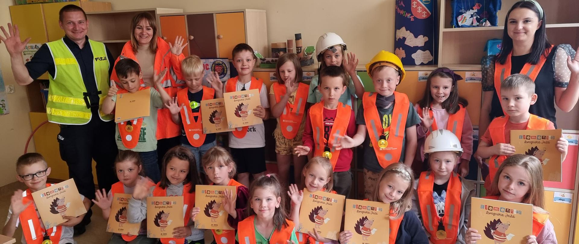 Lekcja bezpieczeństwa - grupa dzieci z książeczkami i elementami odblaskowymi, policjant mundurze, pracownica Oddziału GDDKiA w pomarańczowej kurtce, nauczycielka 