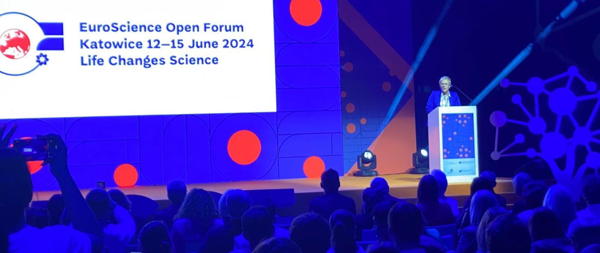 W oświetlonej na niebiesko wypełnionej ludźmi sali za mównicą stoi wiceminister Mrówczyńska i mówi do mikrofonu, za nią na wielkim ekranie napis EuroScience Open Forum 2024 Katowice.