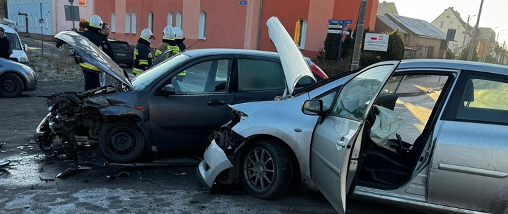 Dzień. Na pierwszym planie stoją dwa samochody osobowe. Otwarte przednie drzwi, uniesione maski. W każdym samochodzie duże uszkodzenia przodów pojazdów. Za samochodami stoi grupa strażaków w białych hełmach i ubraniach specjalnych w kolorze granatowym. 