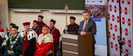 Na krzesłach siedzi kilka osób w ceremonialnych strojach z gronostajowymi kołnierzami, z boku za mównicą stoi minister Wieczorek i mówi do mikrofonu.