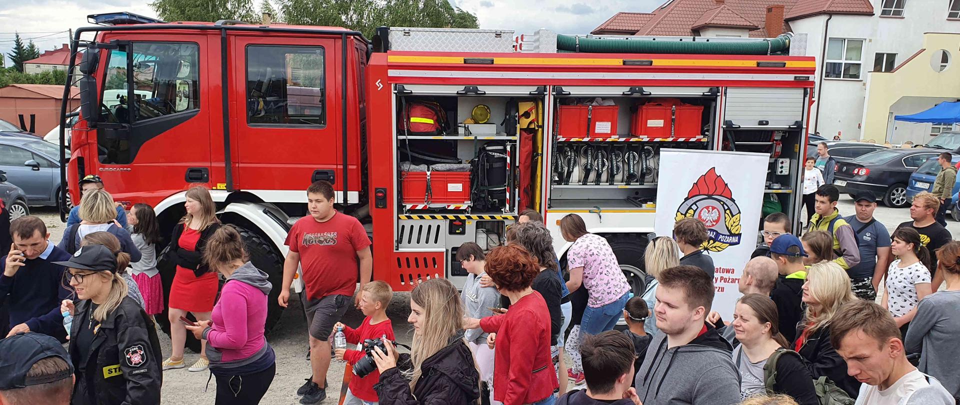 Zdjęcie zrobione w dzień na placu. Na zdjęciu widać dzieci na tle samochodu strażackiego.