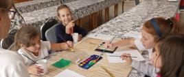 Zdjęcie przedstawia dzieci siedzące przy stole podczas zajęć plastycznych. Dzieci malują pędzlami instrumenty. 