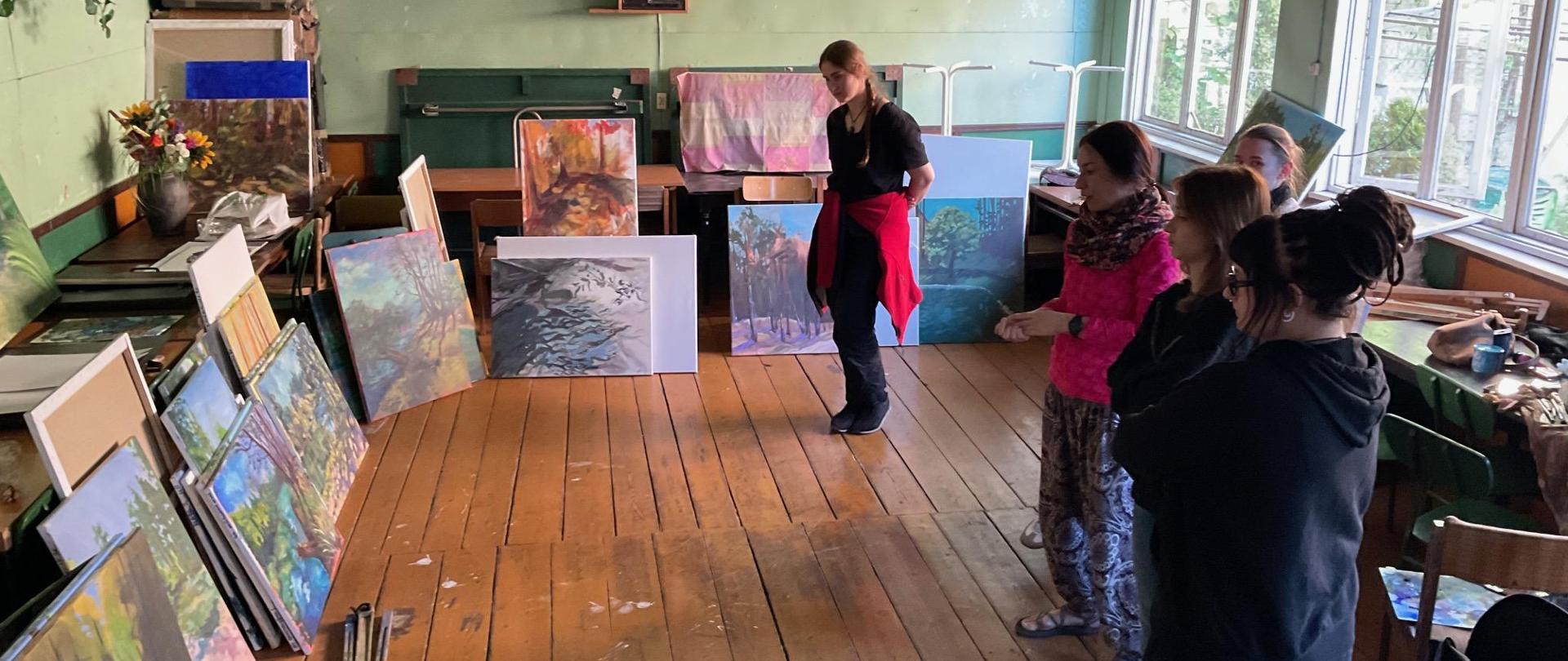 W sali nauczyciel Anna Chmielnik i uczennice stoją przy obrazach malarskich. Nauczycielka omawia prace.
