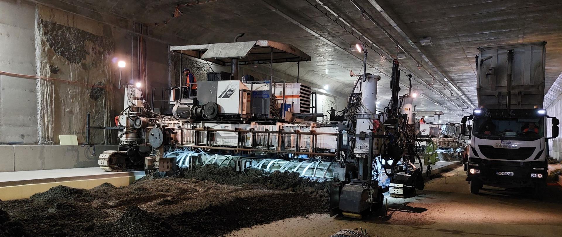 Zdjęcie z wnętrza tunelu na drodze ekspresowej S52. Trwa budowa nawierzchni. Tunelem porusza się maszyna wyrównująca i układająca warstwę nawierzchni.