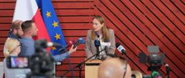 Minister funduszy funduszy i polityki regionalnej Katarzyna Pełczyńska-Nałęcz stoi przy mikrofonach, za jej plecami flagi Polskim i UE, a przed nią dziennikarze
