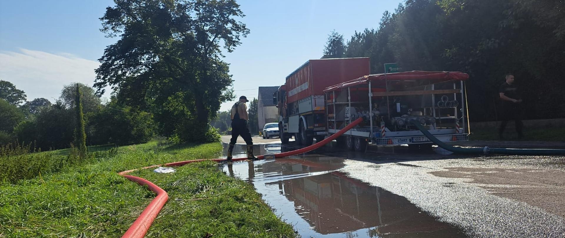 Na zdjęciu duża pompa strażacka z samochodem, obok strażacy i rozlana woda. Pogodnie, słonecznie.