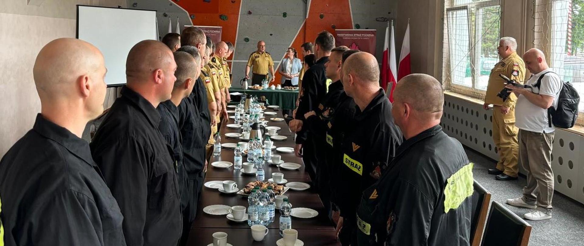 Strażacy zebrani na sali sportowej stoją przy stole w trakcie przywitania Pani Prezydent.