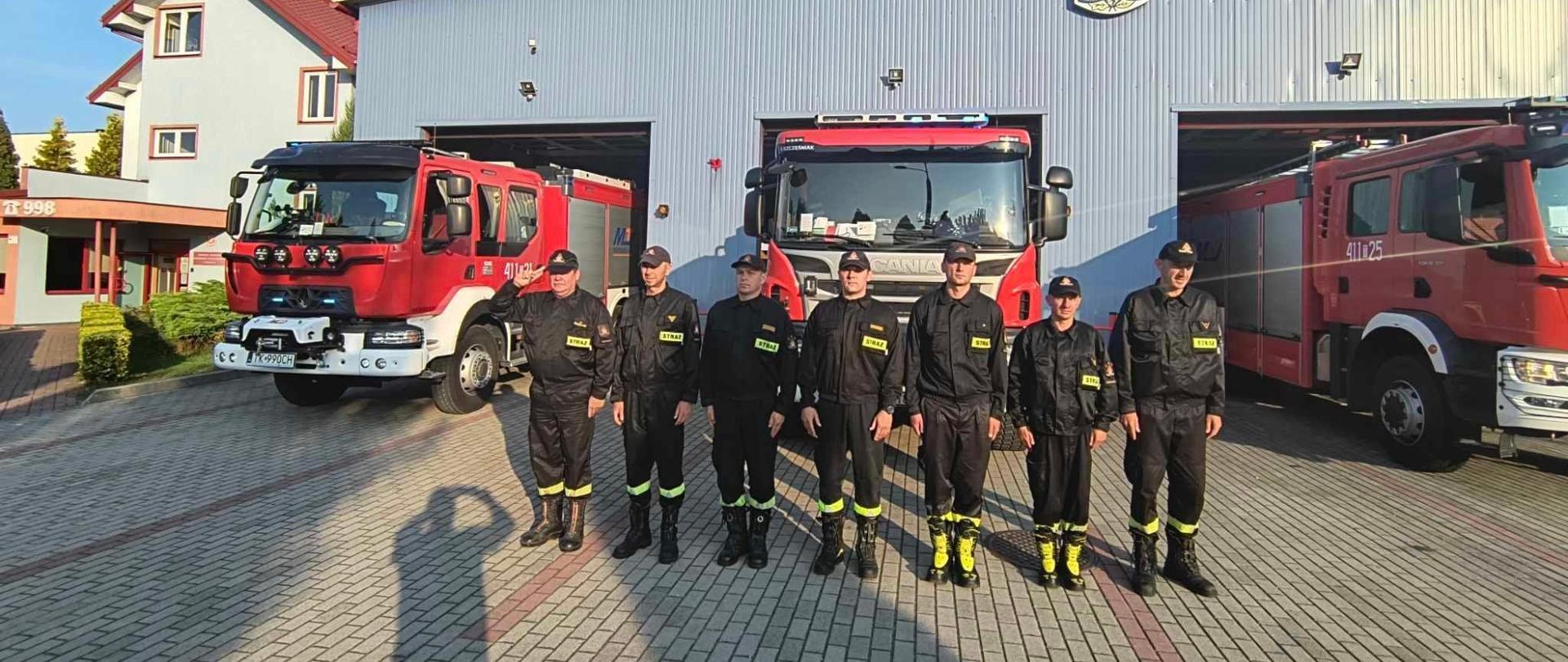 Strażacy na tle pojazdów pożarniczych oddają hołd zmarłym strażakom.