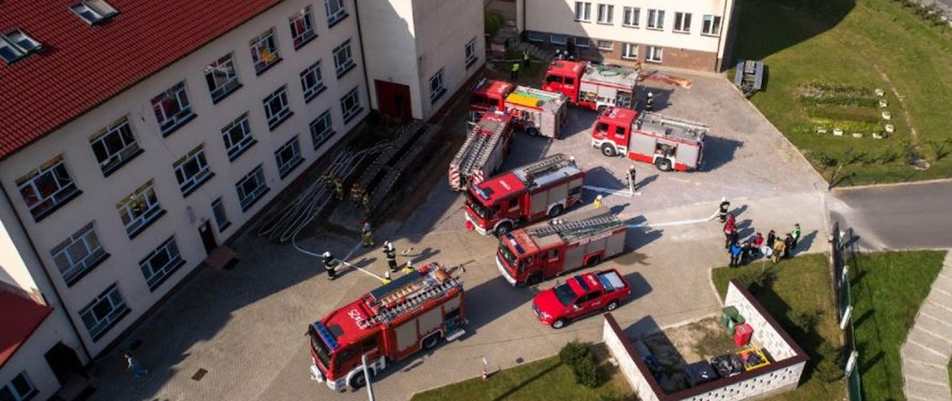 Zdjęcie zrobione z góry, przedstawia plac szkolny a na nim 8 samochodów strażackich. Po lewej widać budynki szkolne przy nich rozłożone węże i strażaków przygotowujących się do rozpoczęcia gaszenia pożaru. Po prawej stronie widać trawni i kawałek ulicy.