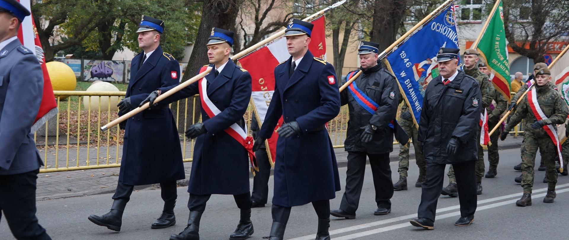 Trzech strażaków maszeruje z pocztem sztandarowym, ubrani na galowo. Za nimi i przed nimi maszerują inne sztandary.