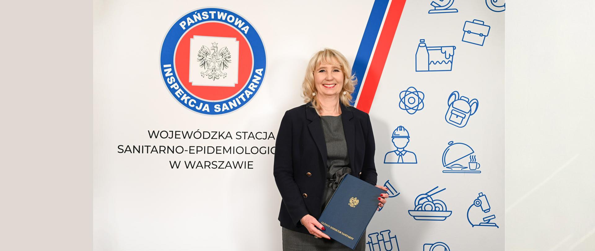 Zastępca Dyrektora WSSE w Warszawie Pani Marzena Woźniak stoi z aktem powołania na tle ścianki z logo Państwowej Inspekcji Sanitarnej.