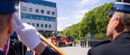 Na zdjęciu podczas powiatowych obchodów dnia strażaka w Gorlicach widoczni od tyłu salutujący i trzymający sztandar strażacy z Ochotniczych Straży Pożarnych a w tle pododdział oraz czerwone samochody pożarnicze. 