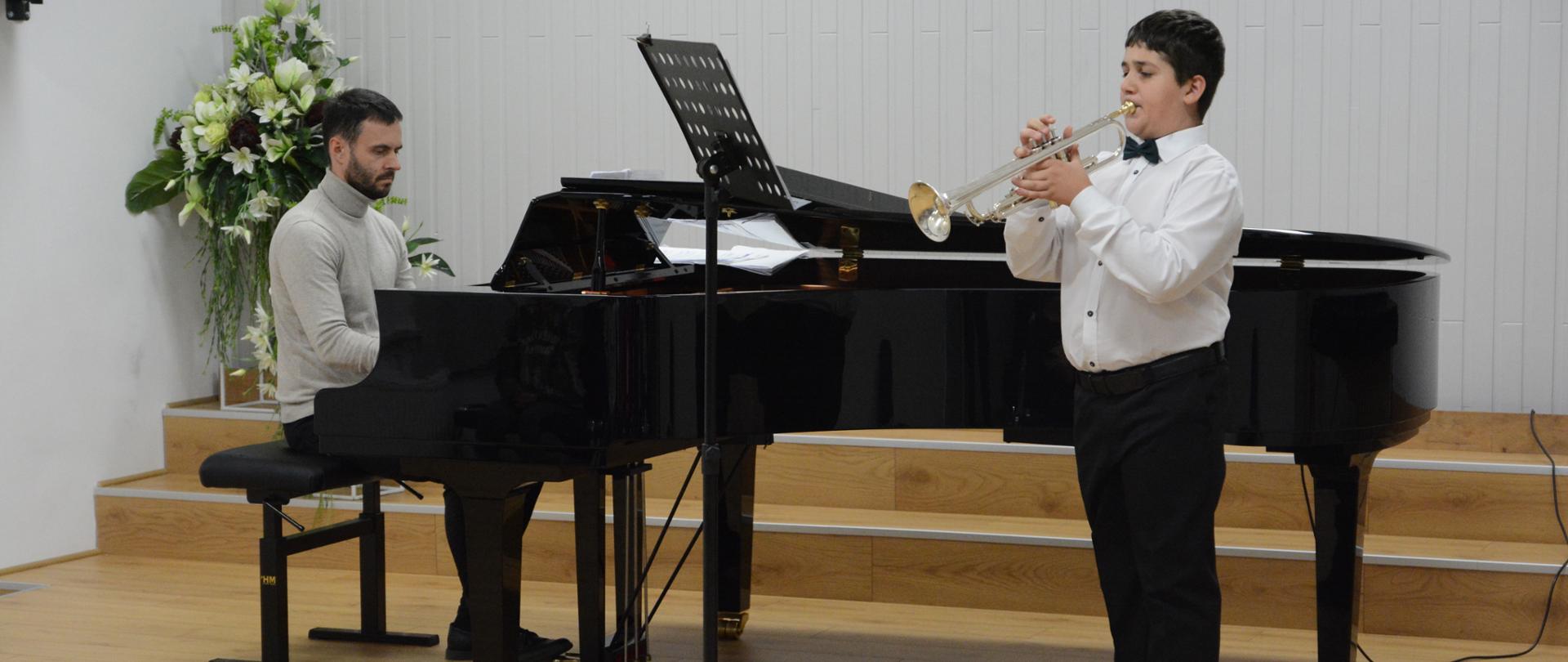 Na zdjęciu utrzymanym w odcieniach bieli i szarośc,i w centralnym punkcie znajduje się fortepian, przy którym pianista akompaniuje uczniowi grającemu na trąbce.