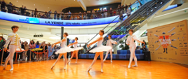 zdjęcie: sześcioro uczniów szkoły baletowe ubranych w białe stroje tańczy klasyczny układ baletowy na scenie w Centrum Handlowym Manufaktura, w tle widoczna publiczność obserwująca występ z parteru i piętra nad sceną, po prawej stronie baner spektaklu "Casanova"