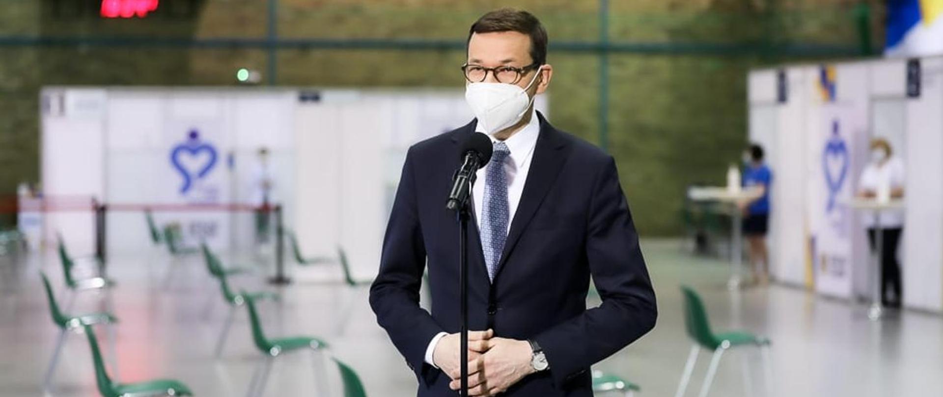Zdjęcie przedstawia premiera - Mateusza Morawieckiego, który znajduje się tuż przed mikrofonem podczas konferencji dot. koronawirusa SARS-CoV-2.