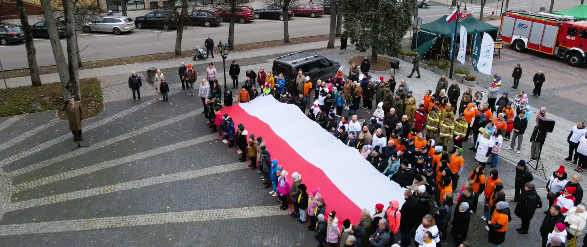 Zdjęcie przedstawia funkcjonariuszy Komendy Powiatowej Państwowej Straży Pożarnej w Gołdapi, którzy o godz. 12:00 na Placu Zwycięstwa w Gołdapi (nieopodal Pomnika Niepodległości) wspólnie z mieszkańcami miasta oraz przedstawicielami władz samorządowych i innych służb zaśpiewali hymn państwowy w ramach akcji „Niepodległa do Hymnu”. Uczestnicy uroczystości trzymają flagę Rzeczypospolitej Polskiej. Z prawej strony fotografii widoczny jest również pojazd pożarniczy. Ujęcie z góry