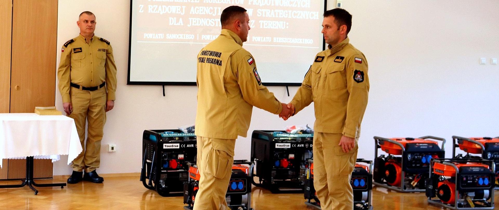 Zdjęcie zrobione w pomieszczeniu w trakcie zbiórki z okazji przekazania agregatów jednostkom Ochotniczych Straży Pożarnych. Na zdjęciu trzech oficerów Państwowej Straży Pożarnej za nimi widać ustawione agregaty prądotwórcze. W pomieszczeniu pod ścianą ustawione są agregaty prądotwórcze nad nimi wyświetlany jest slajd z napisem przekazanie agregatów prądotwórczych z rządowej agencji rezerw strategicznych dla jednostek OSP z terenu powiatu sanockiego, leskiego, bieszczadzkiego.