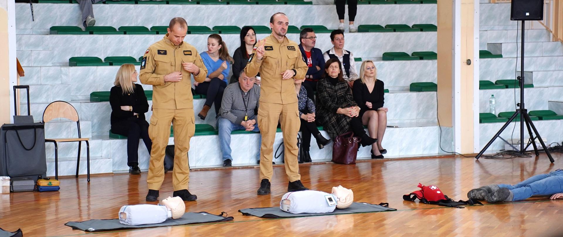 Na zdjęciu 2 strażaków w umundurowaniu służbowym, którzy tłumaczą licealistom zasady udzielania pierwszej pomocy. Przed nimi rozłożone na podłodze manekiny do udzielania pierwszej pomocy. Za ich plecami, na trybunach, grono pedagogiczne.