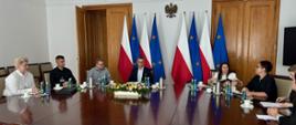Grupa osób siedzi przy owalnym stole, w tle flagi polski i Unii Europejskiej
