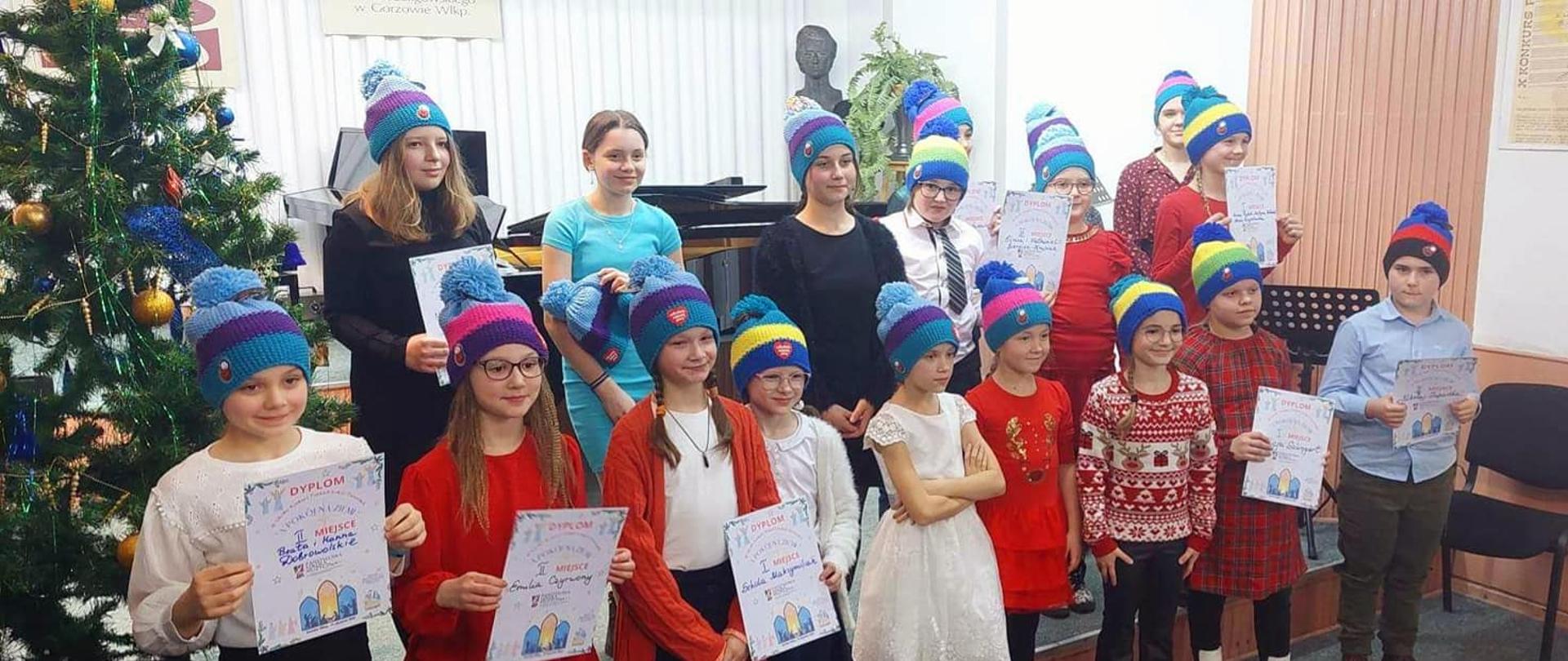Grupa dzieci w czapkach w kolorowe paski, trzymająca dyplomy. Z lewej strony ubrana choinka.