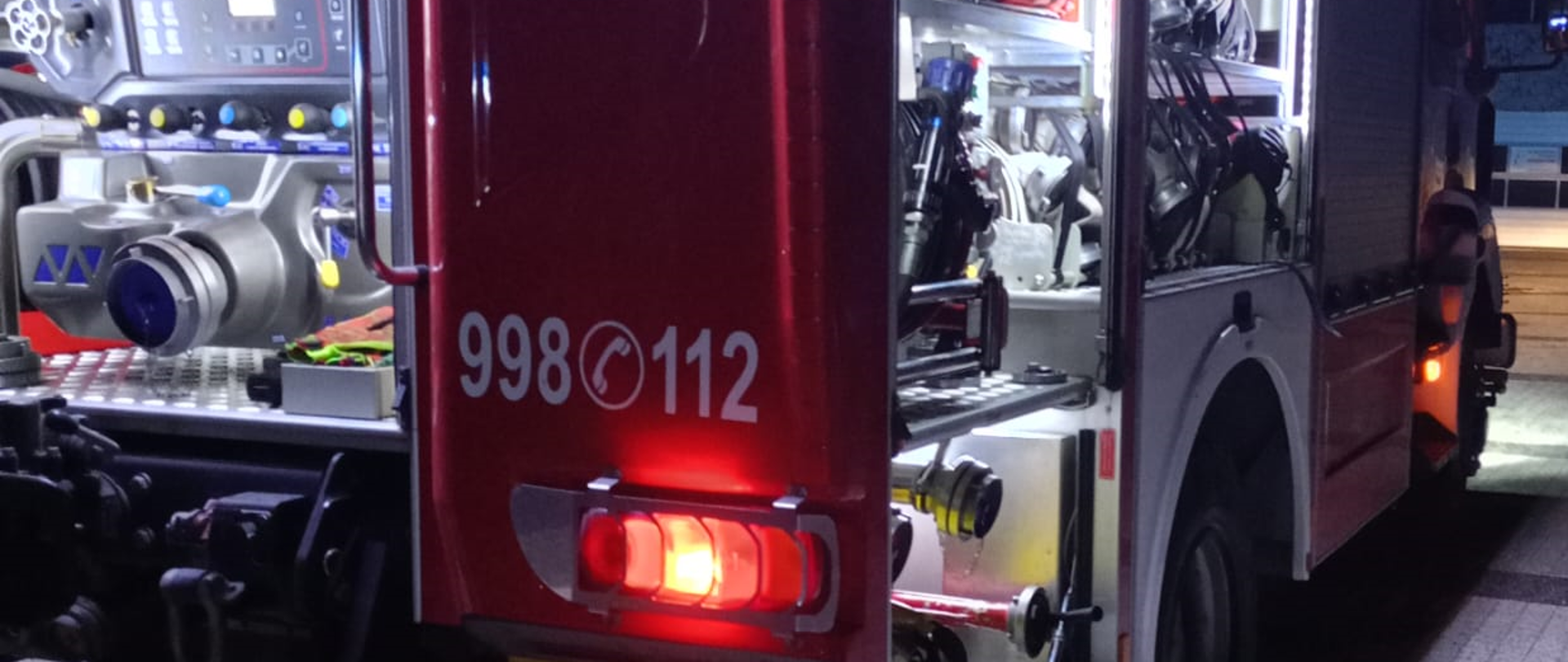 Samochód strażacki stojący tyłem z otwartymi skrytkami. z włączonym oświetleniem 