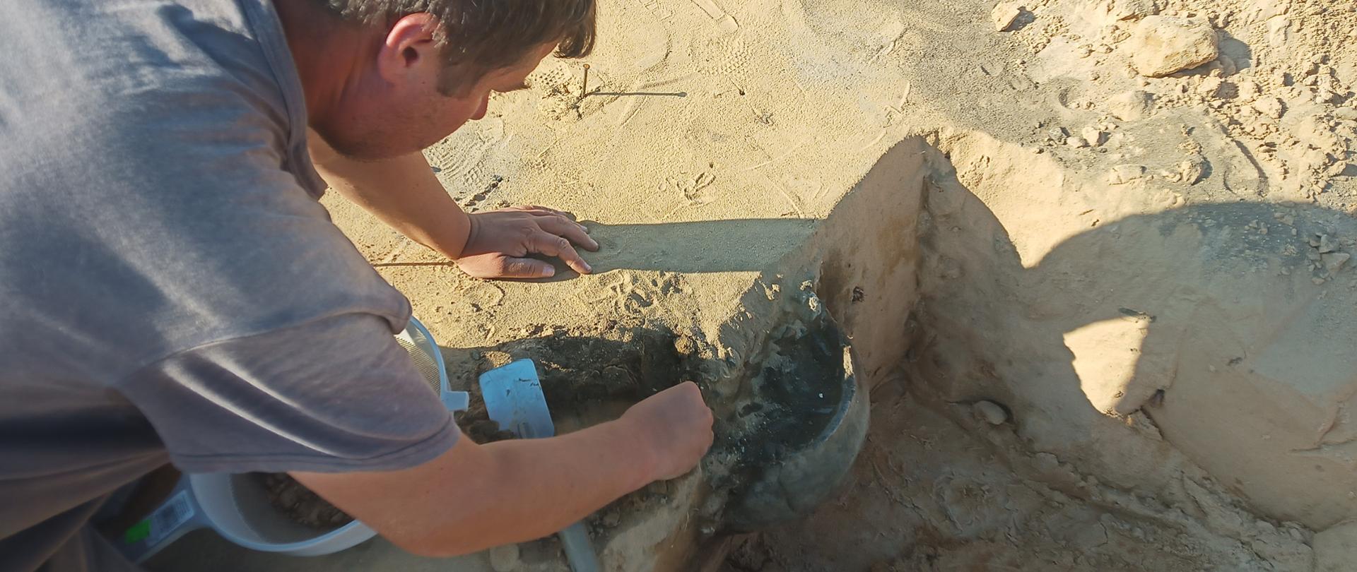 DK73 obwodnica Morawicy - badania archeologiczne - archeolog wykopujący urnę z ziemi, sitko, łopatka, z gruntu wystaje połowa urny 