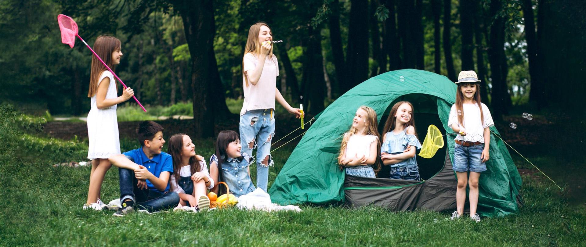 Grupa 8 dzieci bawi się w lesie przy namiocie.