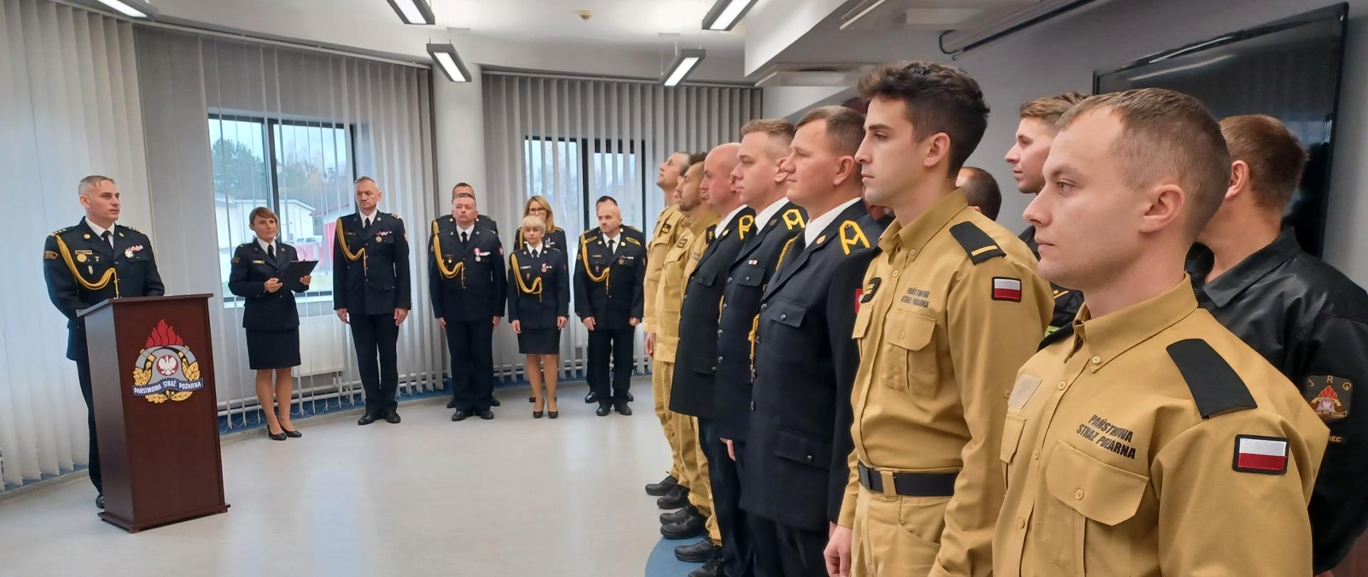 Zdjęcie zrobione w pomieszczeniu strażnicy. Na pierwszym planie po prawej stronie strażacy ustawieni w szeregu, w umundurowaniu służbowym oraz galowym, po lewej komendant powiatowy Państwowej Straży Pożarnej w Lublińcu st. bryg. Janusz Bula stojący za mównicą przemawia do mikrofonu. Na wprost ośmiu strażaków w postawie stojącej w umundurowaniu galowym.