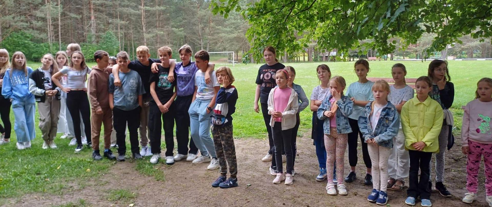 Na zdjęciu duża grupa dzieci. Wszyscy ubrani na sportowo. Zdjęcie wykonane na łonie natury. Dookoła las w oddali bramka do piłki nożnej.
