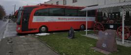 Zdjęcie przedstawia ustawiony przed budynkiem PSP czerwony autobus z napisem Mobilny punkt poboru krwi, z prawej strony pod zadaszeniem stoi zabytkowy wóz strażacki oraz widoczne są 2 tablice pamiątkowe. 