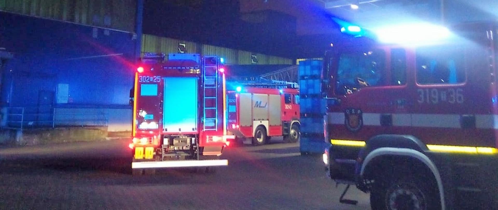 Zdjęcie przedstawia trzy samochody pożarnicze stojące na placu przy hali produkcyjno-magazynowej. W tle hala gdzie doszło do pożaru.